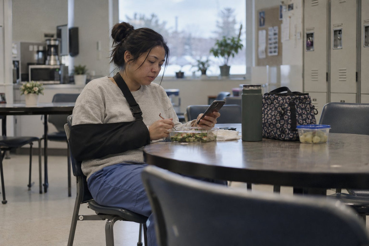 A woman with her arm in a sling eats lunch and checks her phone in a workplace break room on a winter day.