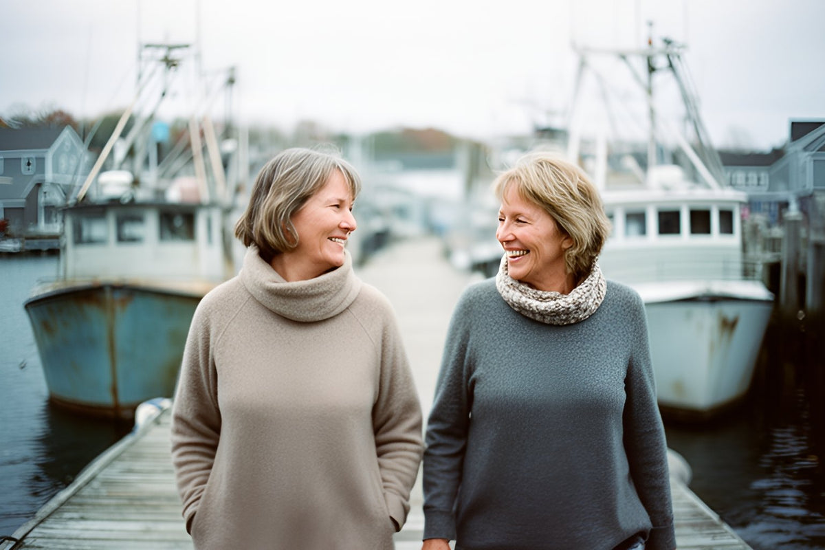 Two women in sweaters walk and laugh together along a harbour wharf, fishing boats tied up behind them.