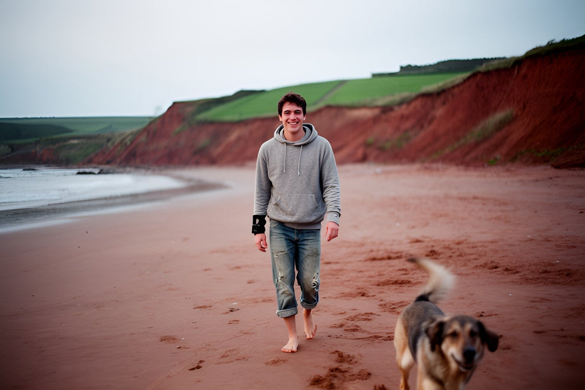 A smiling young man in a grey hoodie and a wrist brace walks barefoot along a red sand beach with his dog, red clay cliffs and green fields behind him.