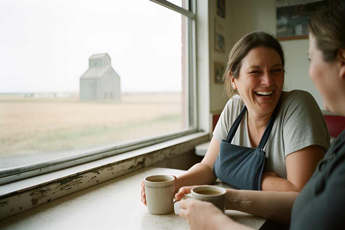 A woman with her arm in a sling laughs with a friend over coffee at a small-town diner window, a grain elevator on the prairie outside.