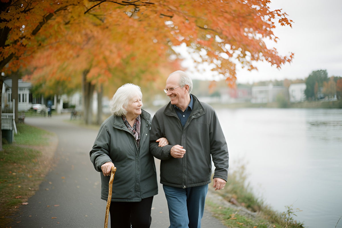 An older woman with a wooden cane walks arm in arm with her husband along a riverside path under orange maple trees.