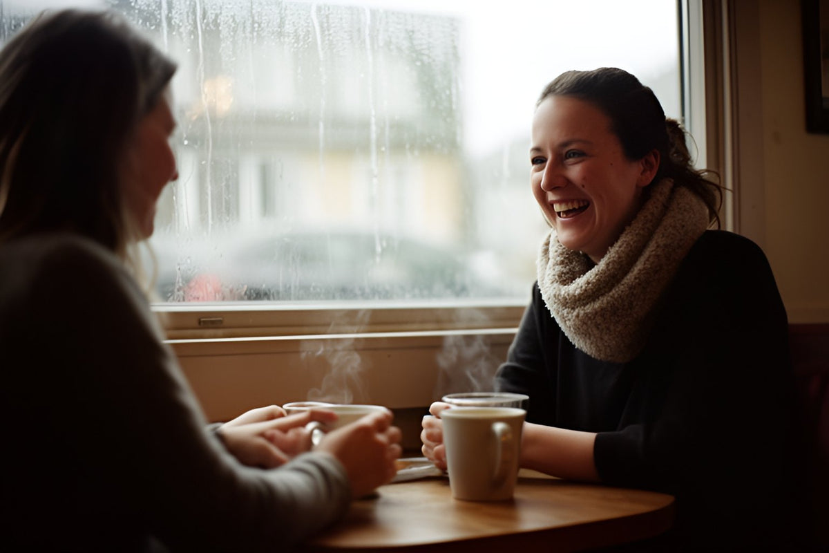 Two friends laugh over mugs of tea at a café table, rain running down the window beside them.